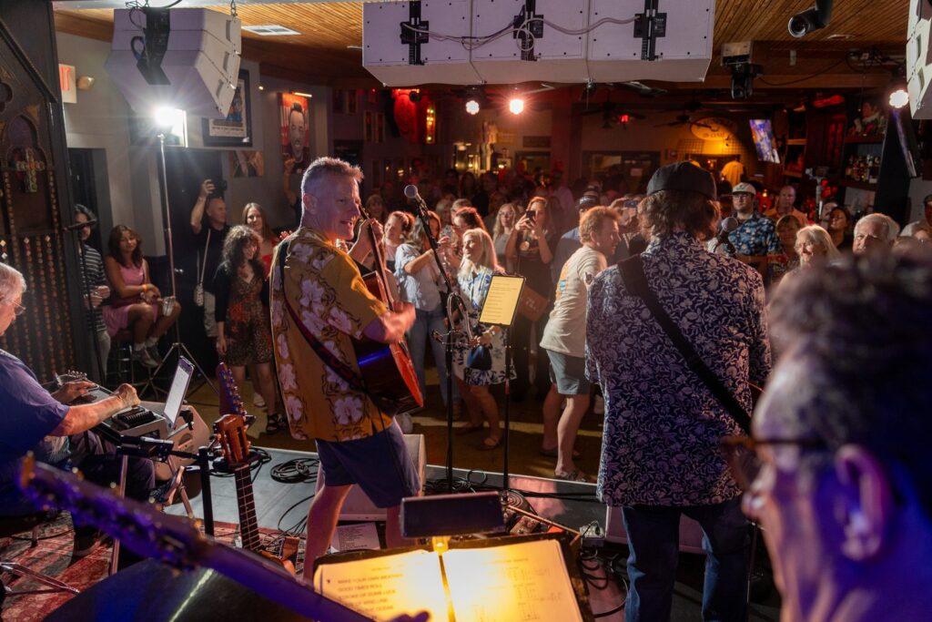 A packed crowd watches a performer in a bold patterned shirt sing at Chickie Wah Wah, another musician in the foreground